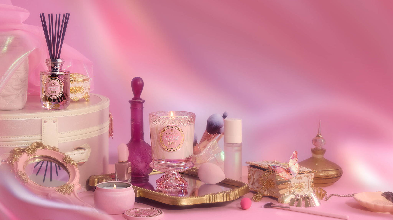 Luxuriöses Interior-Arrangement mit Duftkerze, Parfümflakon, Beauty-Accessoires und goldenen Deko-Elementen auf rosa Hintergrund bei Codeso Living