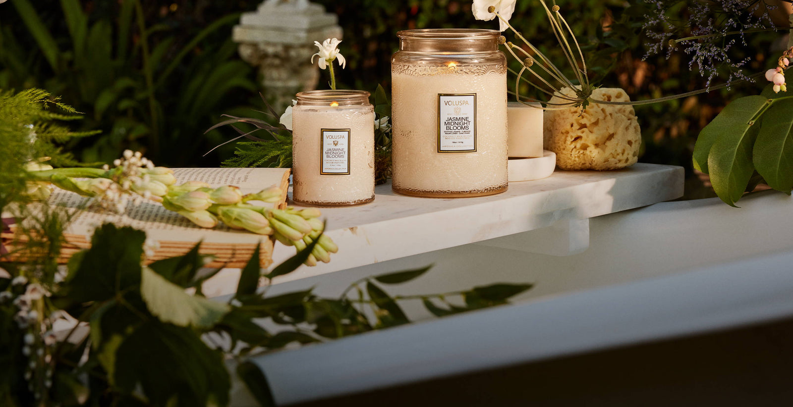 Zwei Voluspa Duftkerzen Jasmine Midnight Blooms auf Marmorplatte, umgeben von Blumen und Grün im Garten, luxuriöses Maximalismus-Interior.