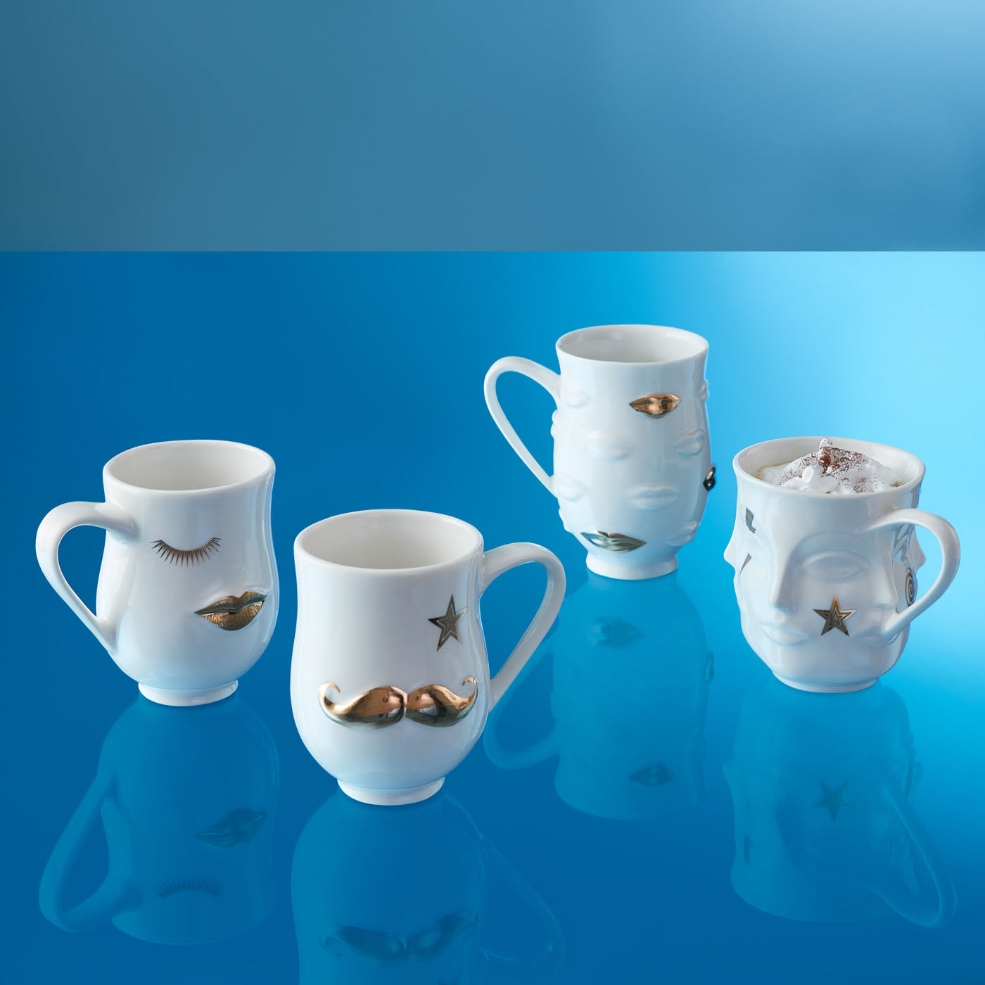 Vier weiße Kaffeebecher von Jonathan Adler mit goldenen Details und Gesichtsmotiven auf blauem Hintergrund
