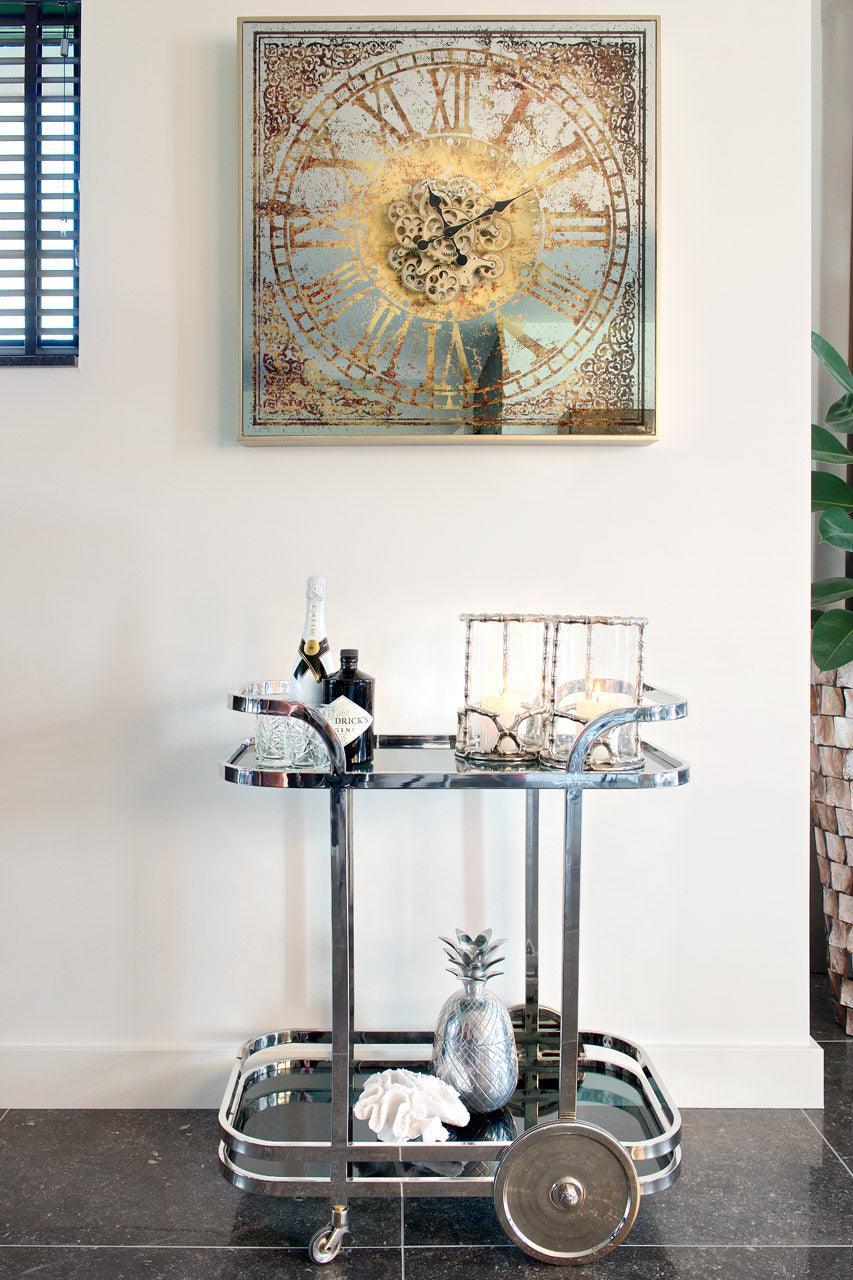 Eleganter Servierwagen aus Chrom mit Sekt, Deko und Gläsern vor großer Vintage-Uhr im Glamour Interior Stil.